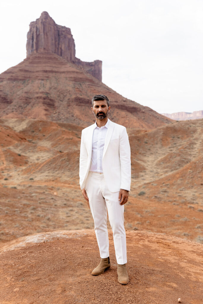 groom standing on red rock wearing white suit