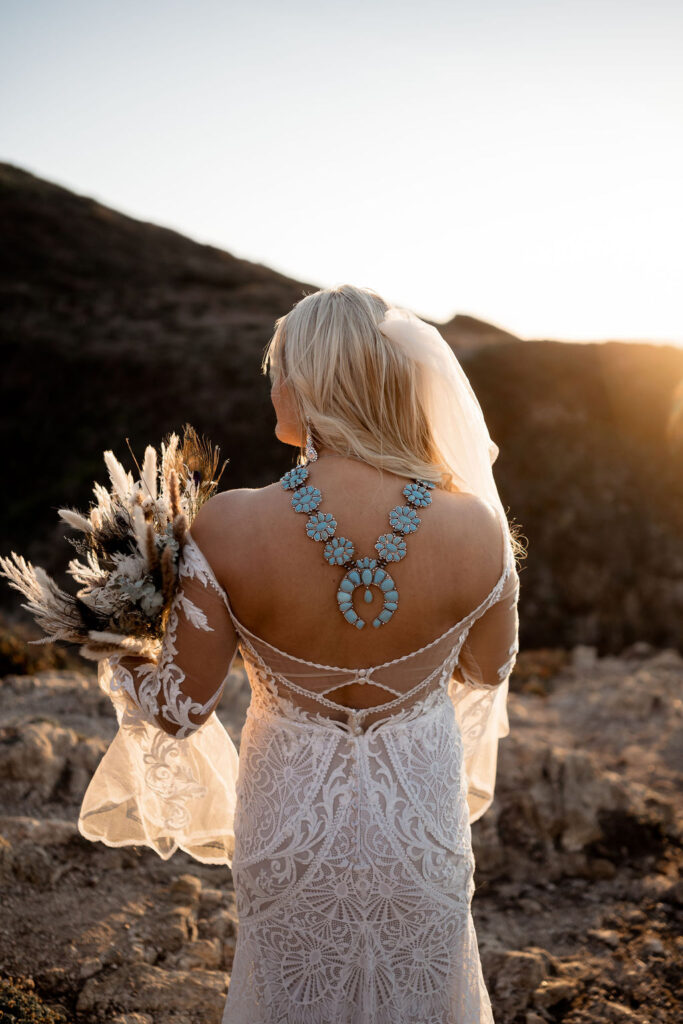 Big Sur elopement photographer captures bride looking away wearing wedding attire holding bridal bouquet and wearing turquoise necklace
