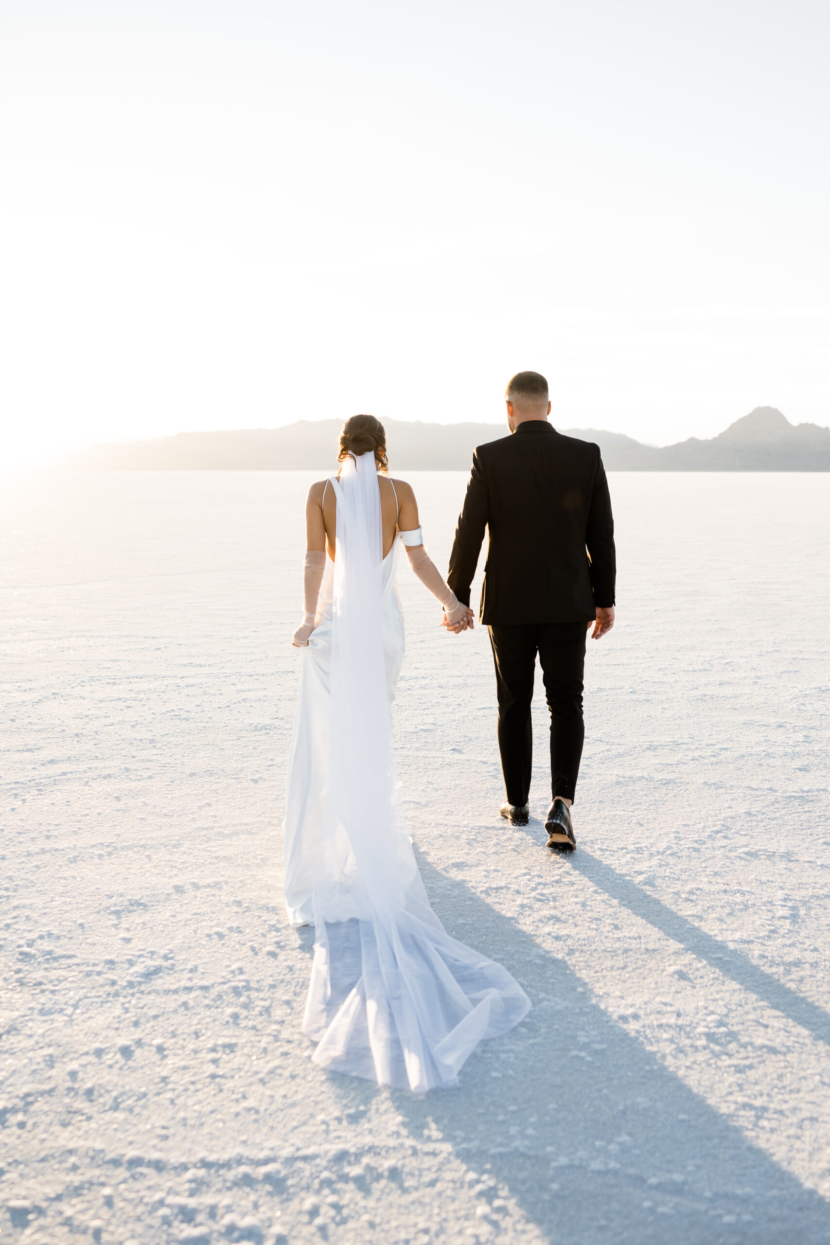 Wide shot of vast Bonneville Salt Flats with married couple in wedding attire holding hands walking into the suset in utah