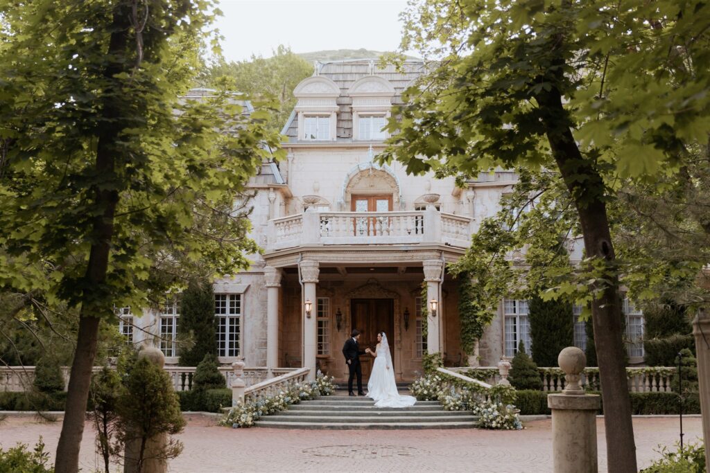 Bride and groom standing together in front of a beautiful, elegant building at La Caille in Utah, surrounded by charming architecture and natural scenery.