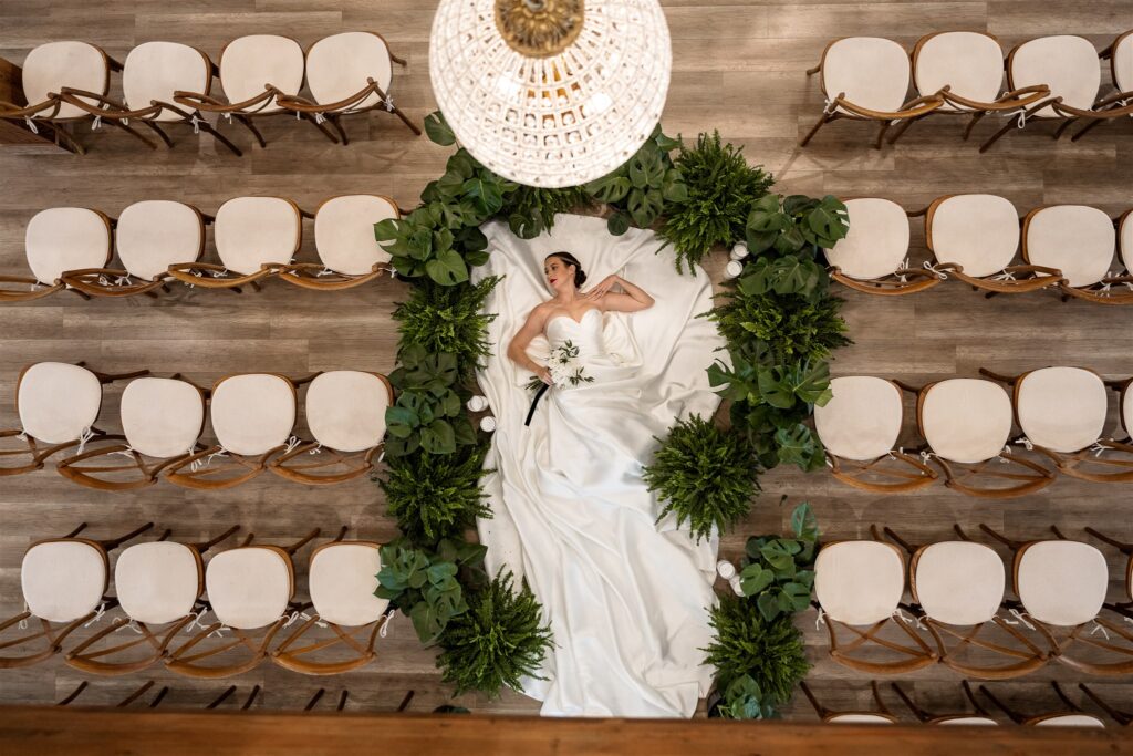 Bride laying on the ground in her wedding dress between the ceremony chair aisle, with her gown elegantly spread out and a dreamy, artistic vibe.