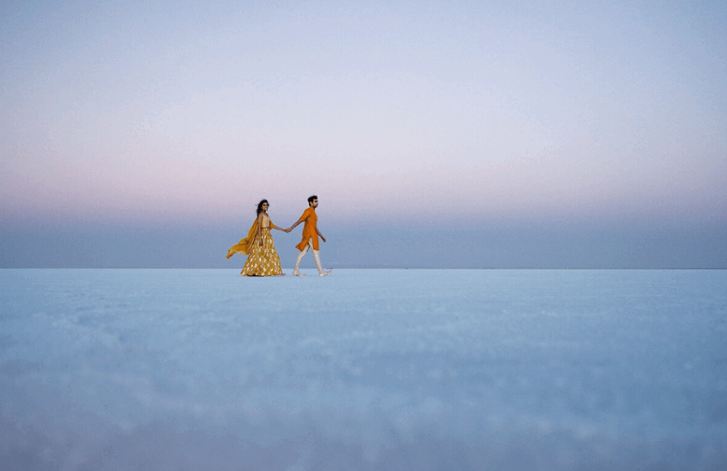 A couple holding hands walks across expansive salt flats at sunset, illuminated by soft golden light against a pastel sky.